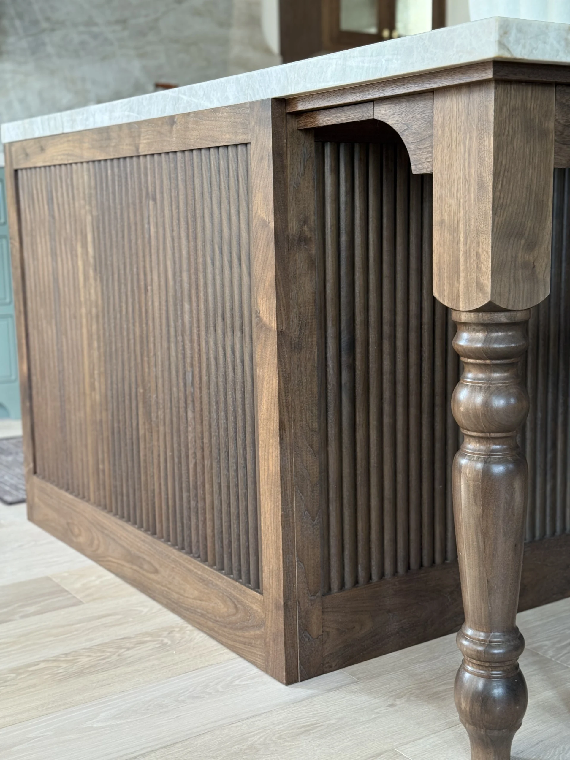 Detail of custom walnut kitchen island with fluted reeded panels, turned leg, and marble countertop